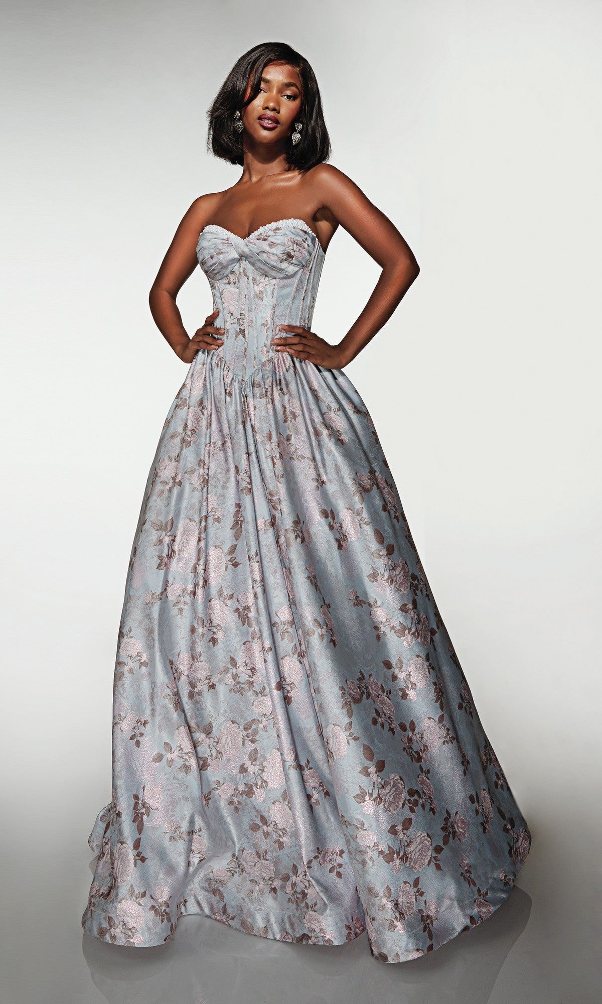 A woman stands with hands on hips, wearing the Alyce 62181 long prom dress—a strapless ball gown with a delicate floral pattern in pale blue and pink tones—set against a plain background.